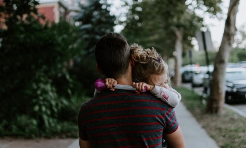 tired little girl rests her head on her father's shoulder as he carries her down the city sidewalk